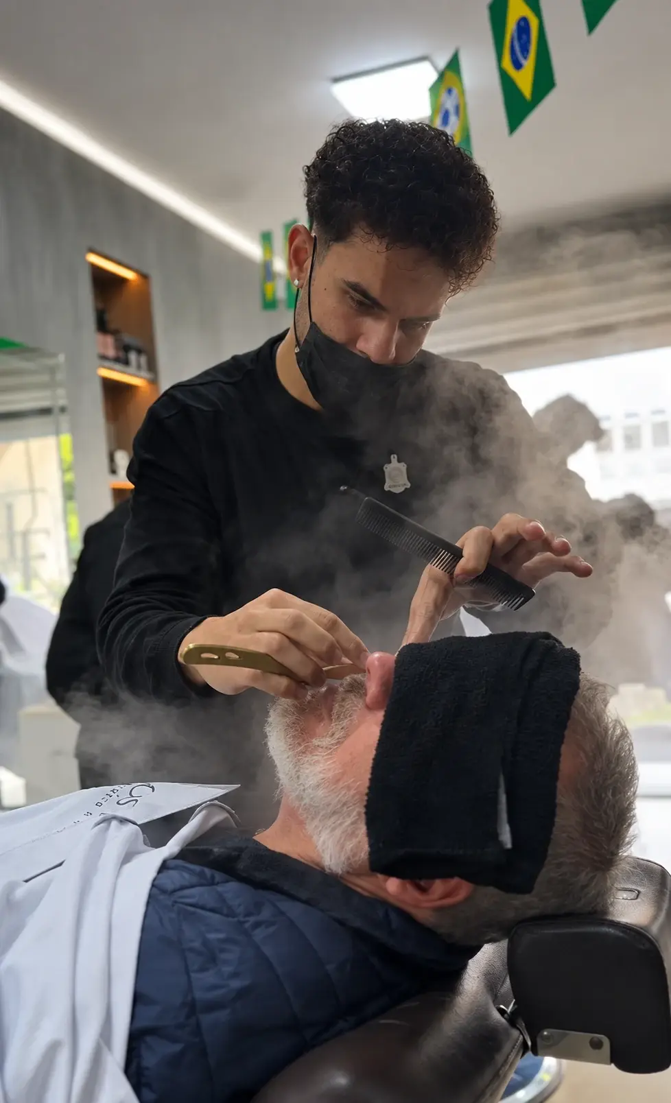 Barbeiro aplicando toalha quente no rosto de um cliente reclinado na cadeira